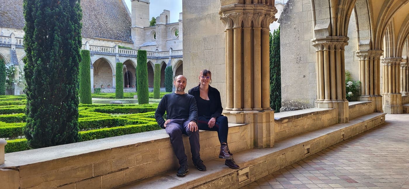 Axelle et Morgan assis sur le muret du cloître de Royaumont, entre deux colonnes, le jardin derrière eux