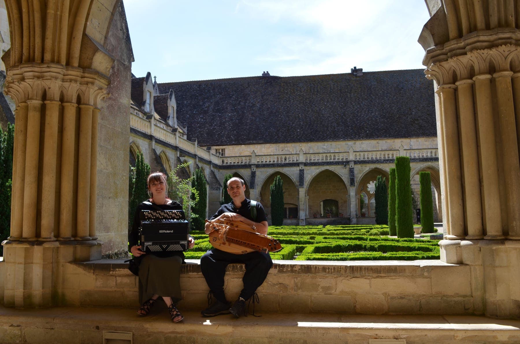 Axelle et Morgan assis sur le banc de pierre d'un cloître, l'accordéon et la vielle à roue sur les genoux, devant les arcades et le jardin taillé